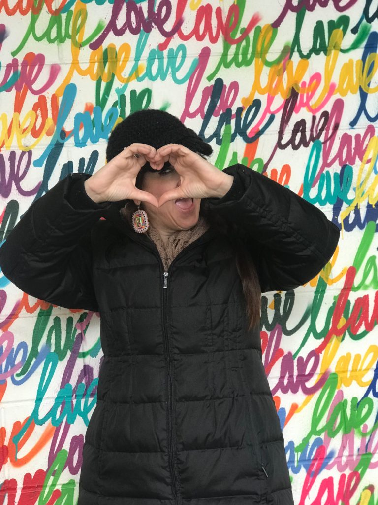 Making the heart sign with hands in front of a Love Mural